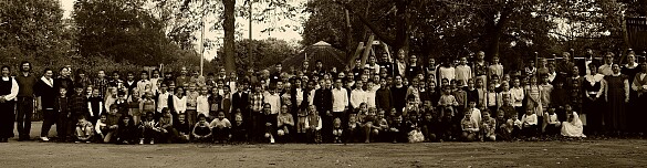 alle Kinder und das ganze Kollegium haben sich auf dem Schulhof versammelt; alle tragen Kleidung, wie man sie vor 100 Jahren getragen hat